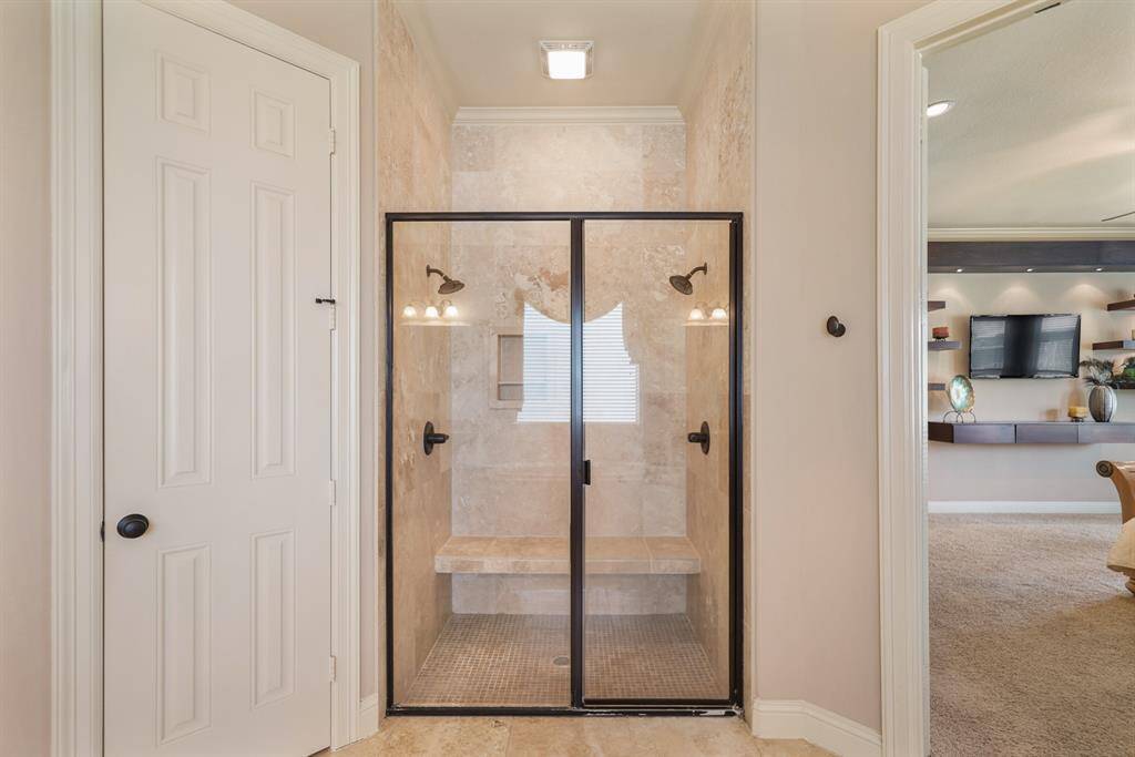 This photo features a luxurious bathroom with a spacious glass-enclosed shower, complete with dual showerheads and a built-in bench. The neutral tile design adds elegance. Adjacent to the shower, you can see a glimpse of a well-lit living area with modern built-in shelving and a wall-mounted TV.