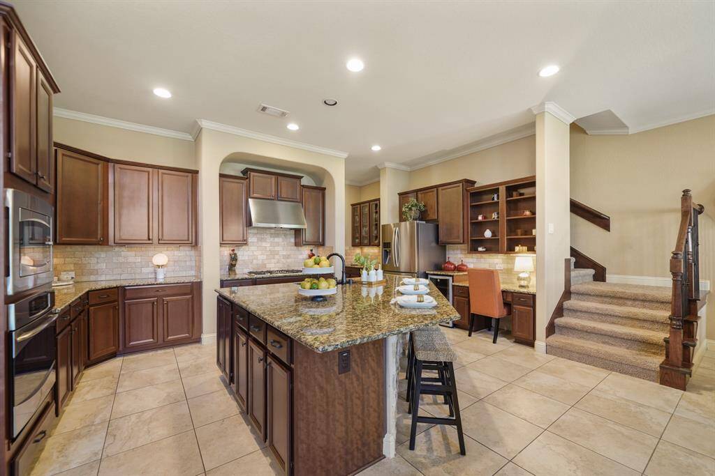This spacious kitchen features elegant granite countertops, an island with seating, and rich wooden cabinetry. It includes modern stainless steel appliances, a cozy desk nook, and easy access to an adjoining staircase. The tile flooring and recessed lighting add a bright, polished look.