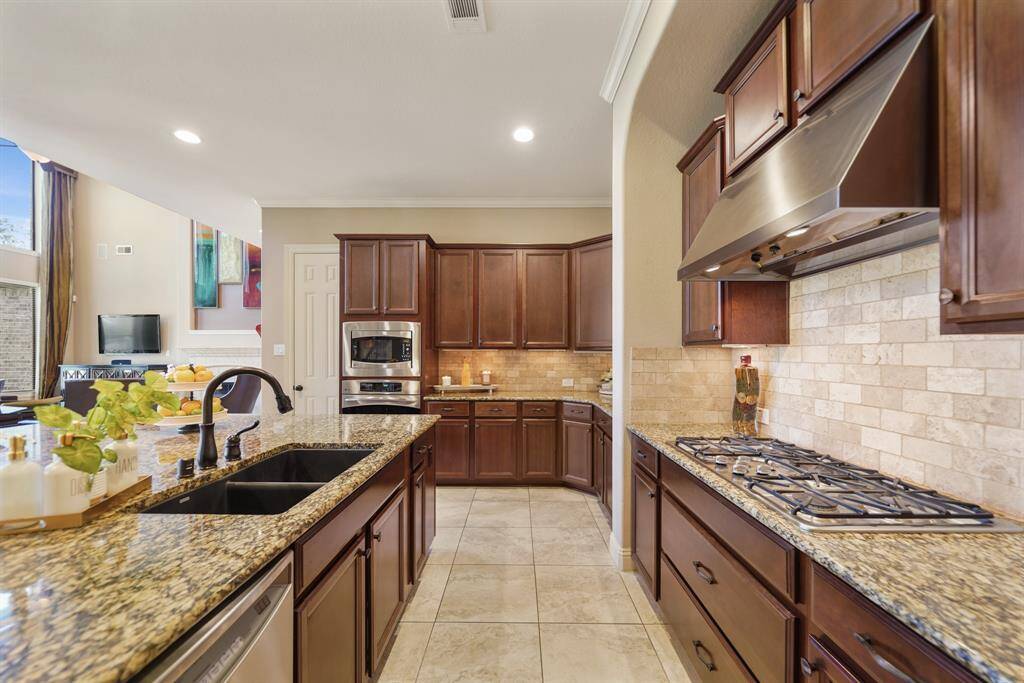 This kitchen features elegant dark wood cabinetry, granite countertops, and a stylish tile backsplash. It includes a modern gas cooktop, stainless steel appliances, and a spacious island with a sink, perfect for cooking and entertaining. Natural light fills the space, highlighting its open and inviting atmosphere.
