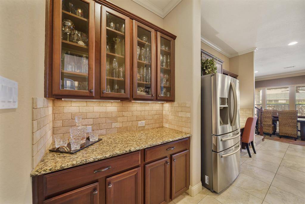 This kitchen features elegant glass-front cabinets, granite countertops, and a stainless steel refrigerator. The space is enhanced by a brick-style backsplash and ample natural light from large windows, creating a warm and inviting atmosphere.