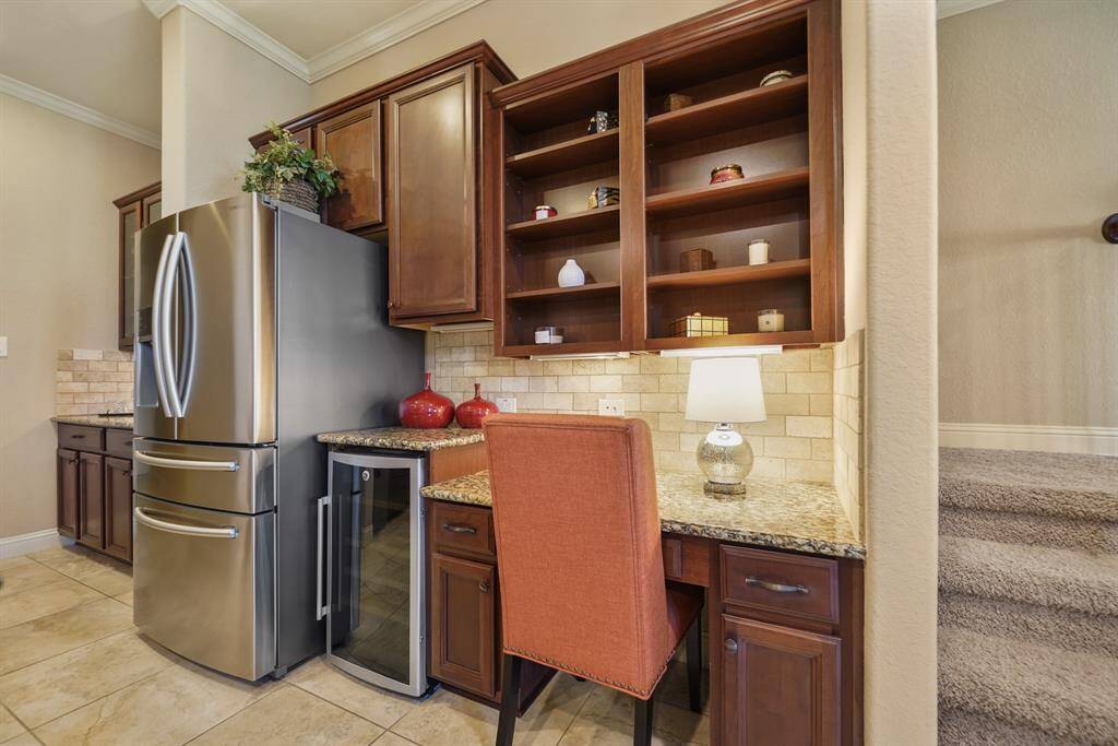 This photo showcases a cozy kitchen nook with built-in shelves and a desk area. It features granite countertops, a stainless steel refrigerator, a wine cooler, and warm wooden cabinetry. The space has tiled flooring and a small lamp, offering a practical and stylish workspace.
