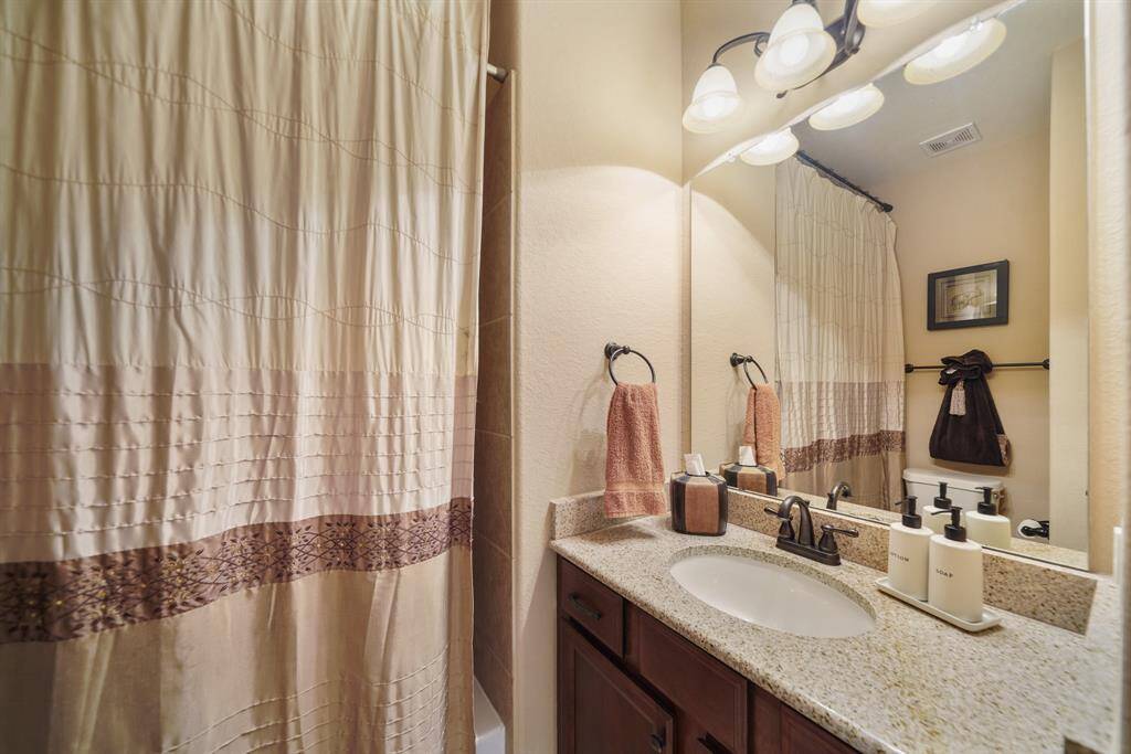 This bathroom features a granite countertop with a single sink, a large mirror, shower/tub combo and elegant lighting.