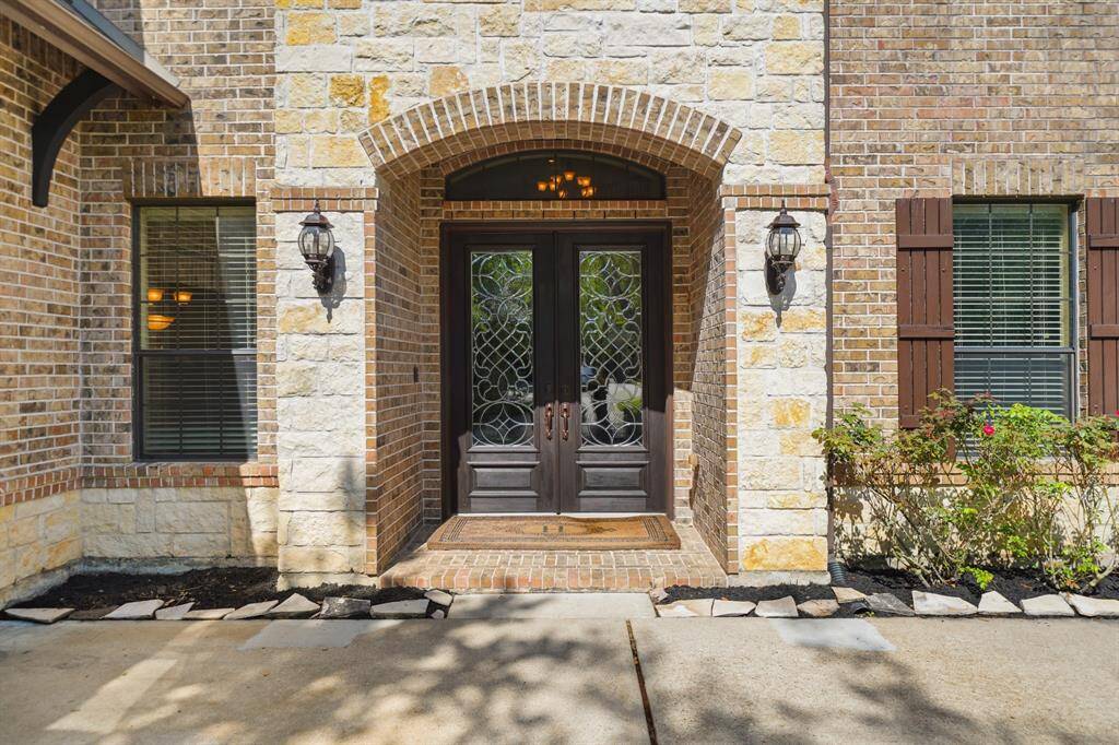 This photo showcases a charming front entryway with a decorative double door, surrounded by a mix of brick and stone exterior. The entry is flanked by two windows with shutters, and there's tasteful landscaping and pathway lighting.