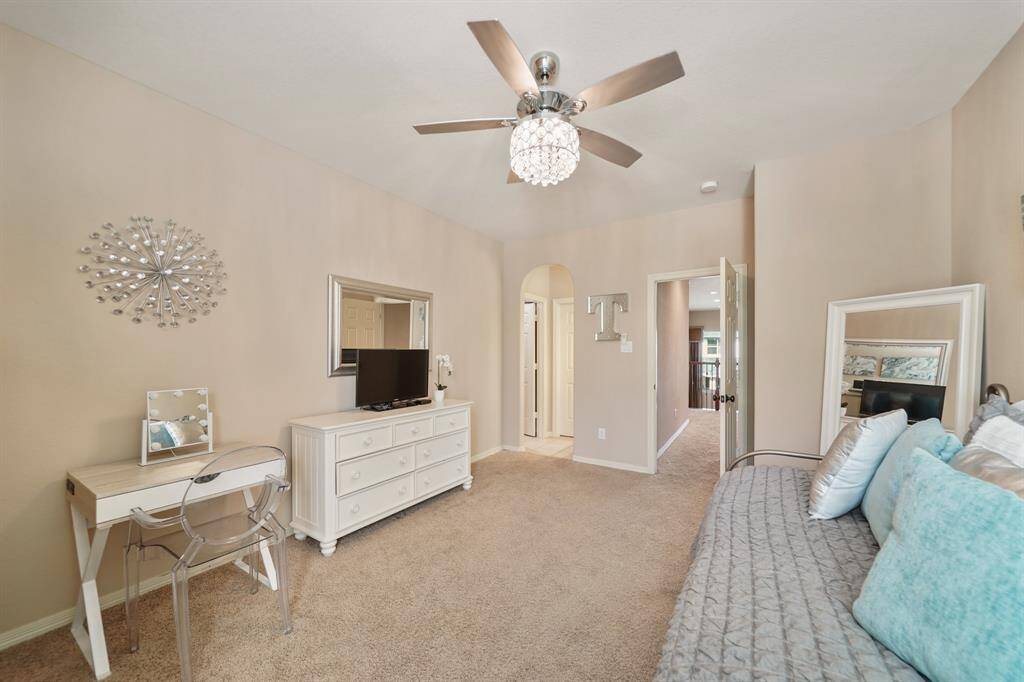 This room features beige walls, plush carpet, and a stylish ceiling fan with a chandelier. The space is bright and airy, with an open doorway leading to a hallway.