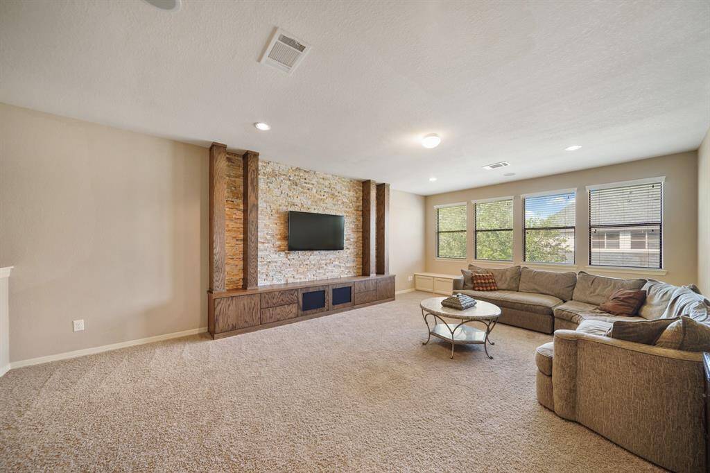 This spacious living room features a cozy carpet, a large sectional sofa, and a stone accent wall with a mounted TV. Natural light streams through multiple windows, creating a bright and inviting atmosphere.