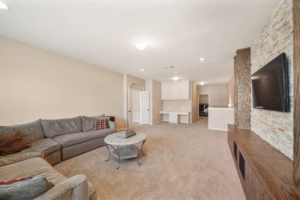 This gameroom features a cozy, carpeted space with a large sectional sofa, wall-mounted TV, stone accent wall, and built-in wooden shelving. The neutral paint and recessed lighting create a welcoming atmosphere, with an open layout leading to adjacent rooms.