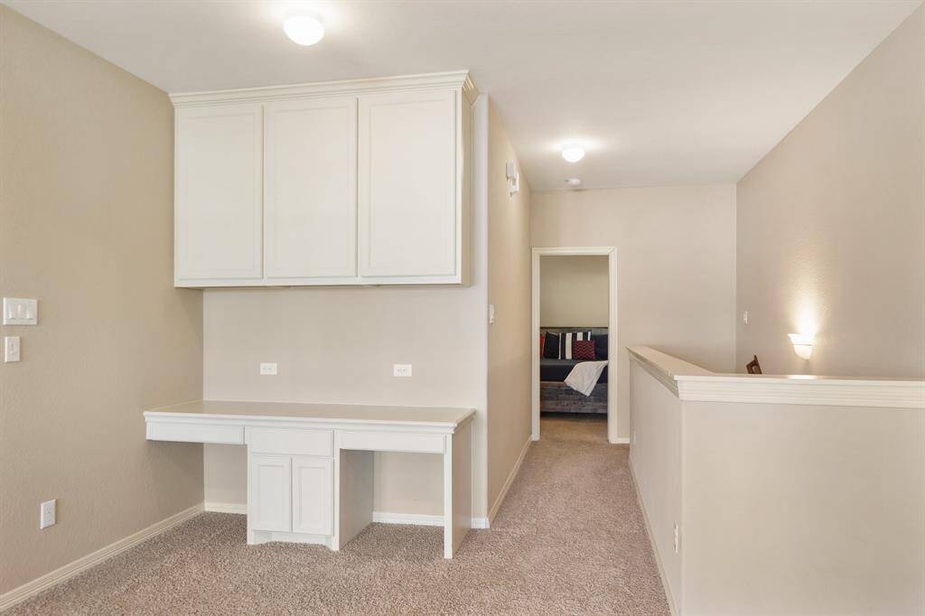 This photo shows a hallway with beige walls and carpet, featuring built-in cabinetry and a desk area. It leads to a cozy room with a glimpse of furniture and decor.