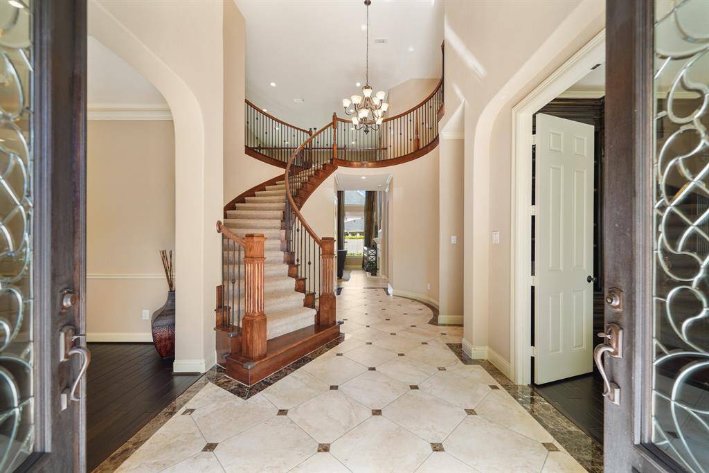 This elegant entryway features a grand curved staircase, high ceilings, and a chandelier. The space is bright with tile flooring and dark wood accents, offering a welcoming and sophisticated first impression.