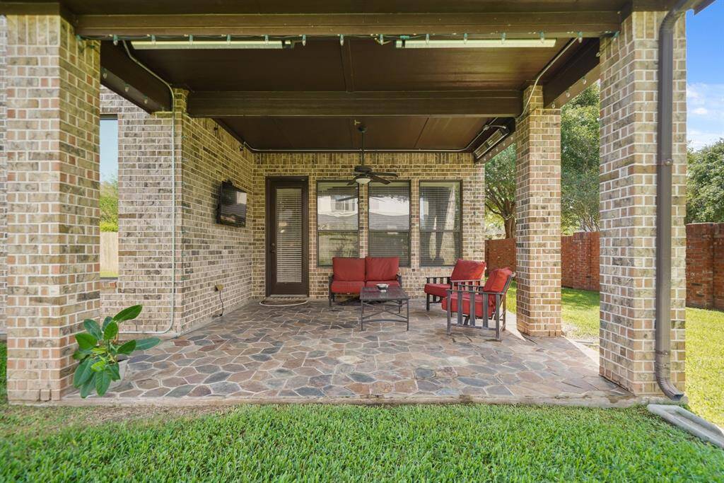 This photo showcases a covered patio with brick columns and a stone floor, furnished with red cushioned seating and a coffee table. There's a wall-mounted TV, ceiling fan, and a view of a well-maintained lawn, perfect for relaxing or entertaining.