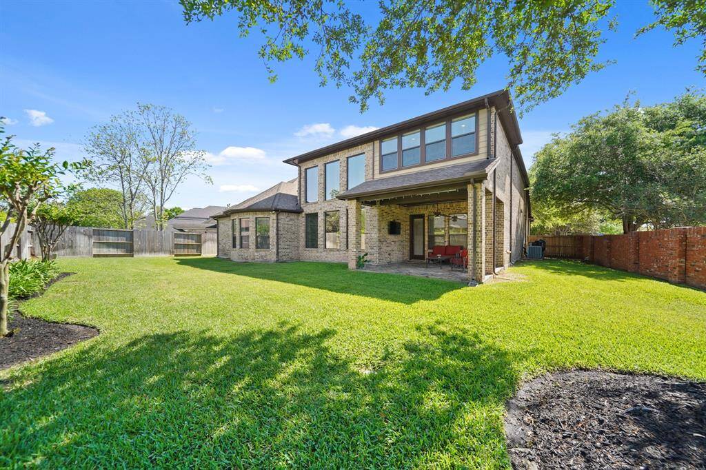 This photo showcases a spacious backyard with lush green grass and a well-maintained brick two-story home. There's a covered patio area perfect for outdoor relaxation, surrounded by a wooden privacy fence and mature trees.