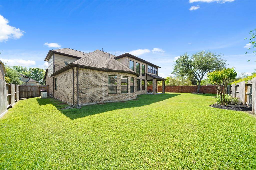 The photo showcases a spacious backyard with a well-maintained lawn, bordered by a wooden fence. The brick house features large windows and a covered patio area, offering plenty of natural light and outdoor relaxation space. Ideal for families and entertaining.