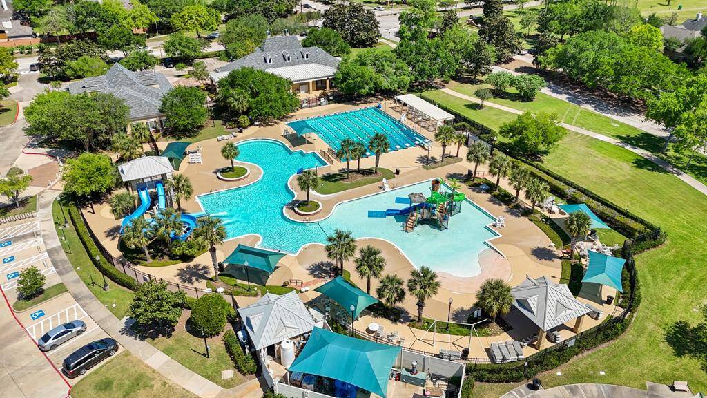 This photo showcases a community recreational area with a large, uniquely shaped pool, featuring water slides and a separate lap pool. There are shaded seating areas, lush landscaping, and nearby parking. The setting is surrounded by greenery, providing a serene and inviting environment for families.