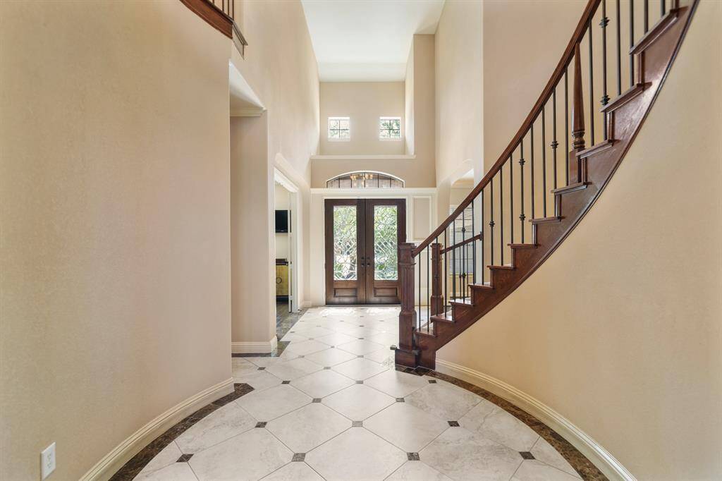 This elegant entryway features a grand, curved staircase with wooden railings, high ceilings, and abundant natural light from the upper windows. The tiled floor leads to a double front door with decorative glass, creating a welcoming and spacious atmosphere.