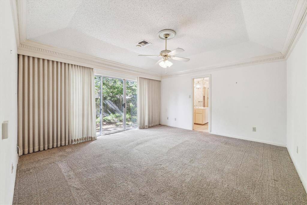 Primary bedroom with neutral carpet, sliding glass doors to a garden, and an attached bathroom. Features a ceiling fan and crown molding for added elegance.