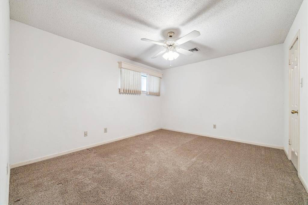 Secondary bedroom features neutral carpeting, white walls, a ceiling fan, and a single window with vertical blinds, offering a simple and clean appearance.