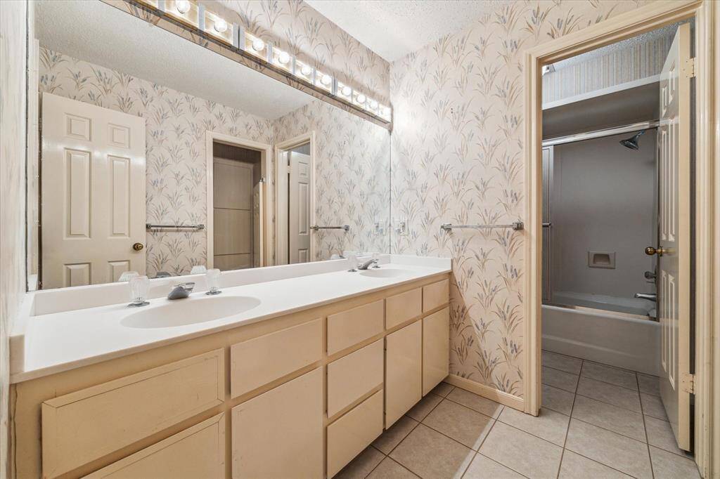 The secondary bathroom that services bedroom 2 and 3, features dual sinks with plenty of cabinet storage, tiled flooring, and a large mirror. The wallpaper adds a classic touch, and there's a bathtub and shower combination in an adjacent space.