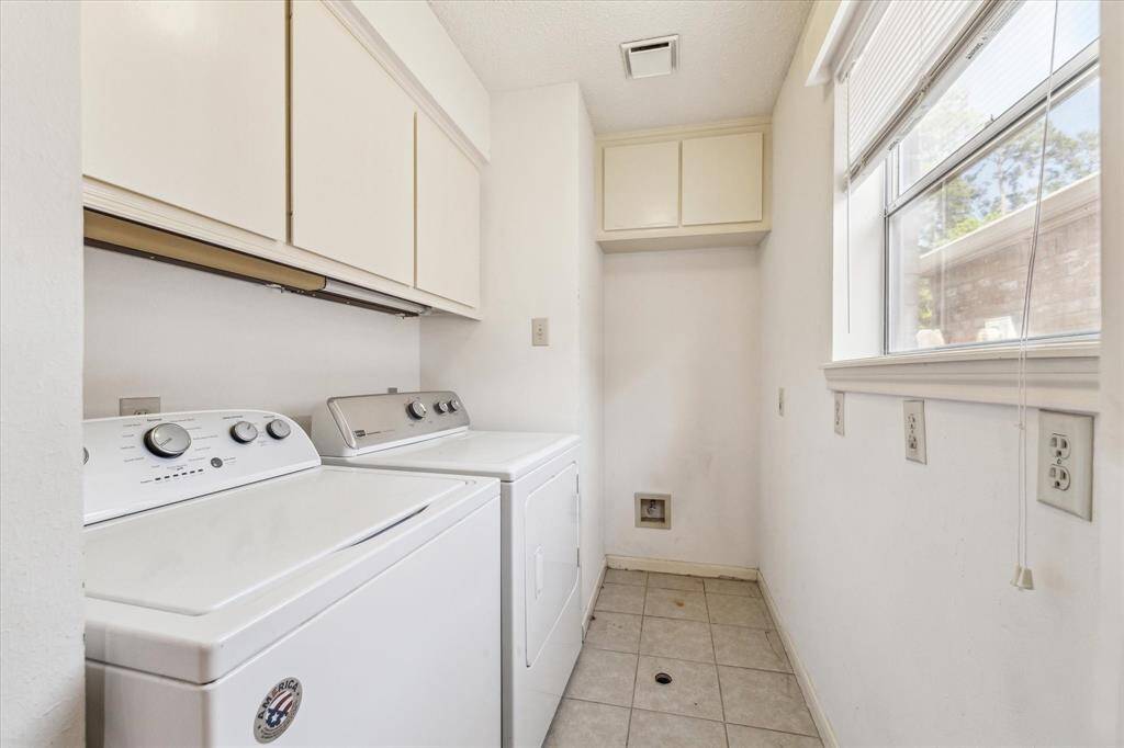 This photo shows a bright, compact laundry room featuring a washer and dryer, upper cabinets for storage, tiled flooring, and a large window allowing natural light.