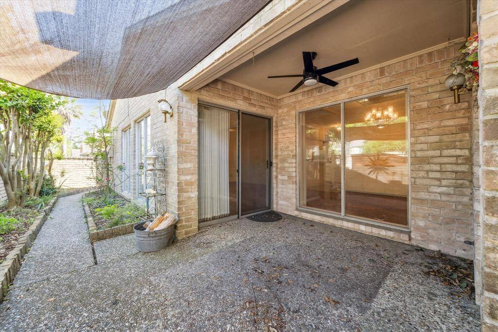 This photo shows a cozy patio area with a ceiling fan and sliding glass doors, leading to a well-maintained garden path. The space is shaded and features brick walls, providing a comfortable outdoor living area.