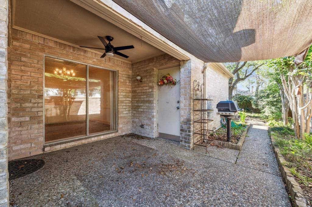 This photo showcases a cozy patio area with brick walls, featuring a ceiling fan and sliding glass doors. There's a small garden path leading to a lush, green backyard, complete with a grill and partial overhead shade. Perfect for outdoor relaxation. The outdoor closet provides space for those gardening and barbeque items for convenience.  There is another outdoor storage on the side of the home with shelving.