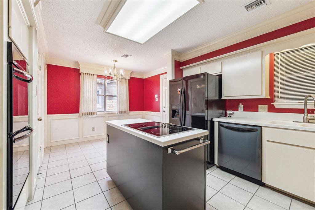 Bright kitchen with striking red walls and white wainscoting. Features include a central island with cooktop, black appliances, ample cabinetry, and a chandelier over the dining area. Tile flooring completes the space.