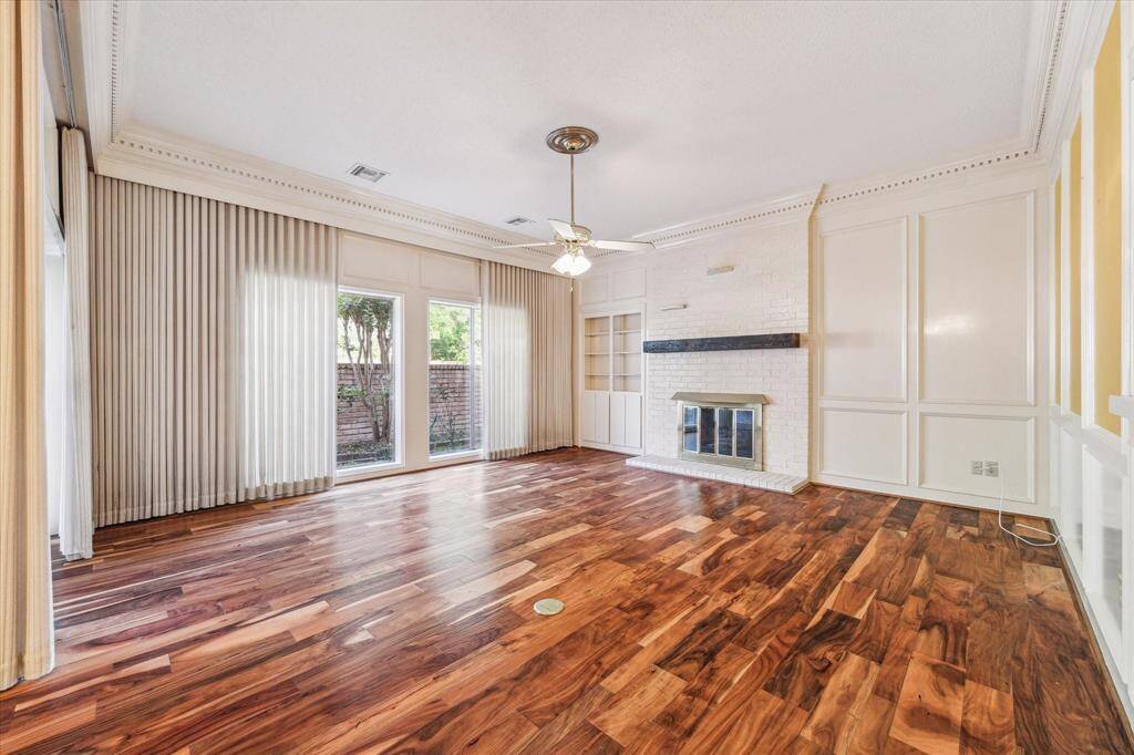 This inviting living room features rich hardwood floors, a cozy brick fireplace, and large windows with sliding doors that offer plenty of natural light and a view of the backyard. Elegant paneling and a ceiling fan add charm and comfort.