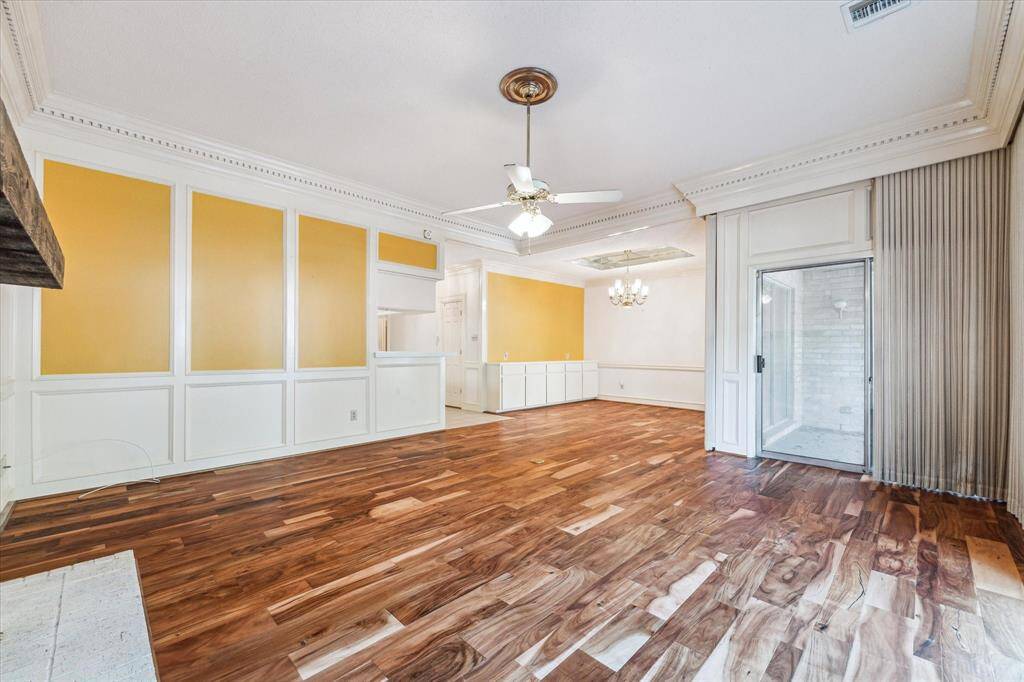 This spacious living and dining area features rich hardwood floors, elegant crown molding, and a ceiling fan. It has a sliding glass door leading to an outdoor space and a unique accent wall with white and mustard panels. A chandelier adds charm to the adjoining dining area.