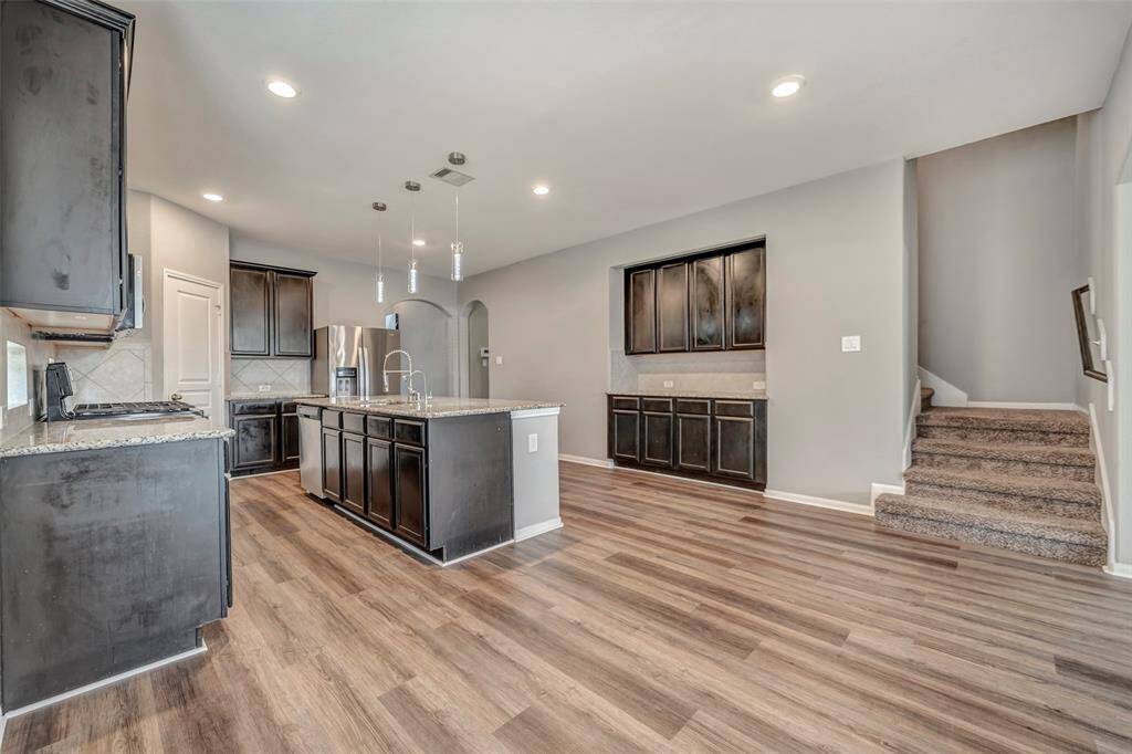 This spacious kitchen features modern dark cabinetry, granite countertops, and stainless steel appliances. It includes a large island with a sink and hanging pendant lights. The open layout extends to a living area with wood-style flooring and a carpeted staircase leading upstairs, creating a warm and inviting atmosphere.
