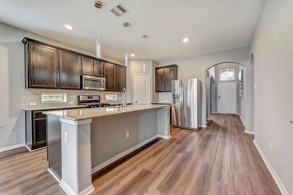 This modern kitchen features sleek dark cabinets, stainless steel appliances, and a spacious island with granite countertops. The open layout flows into the entryway, highlighted by wood flooring and contemporary lighting.