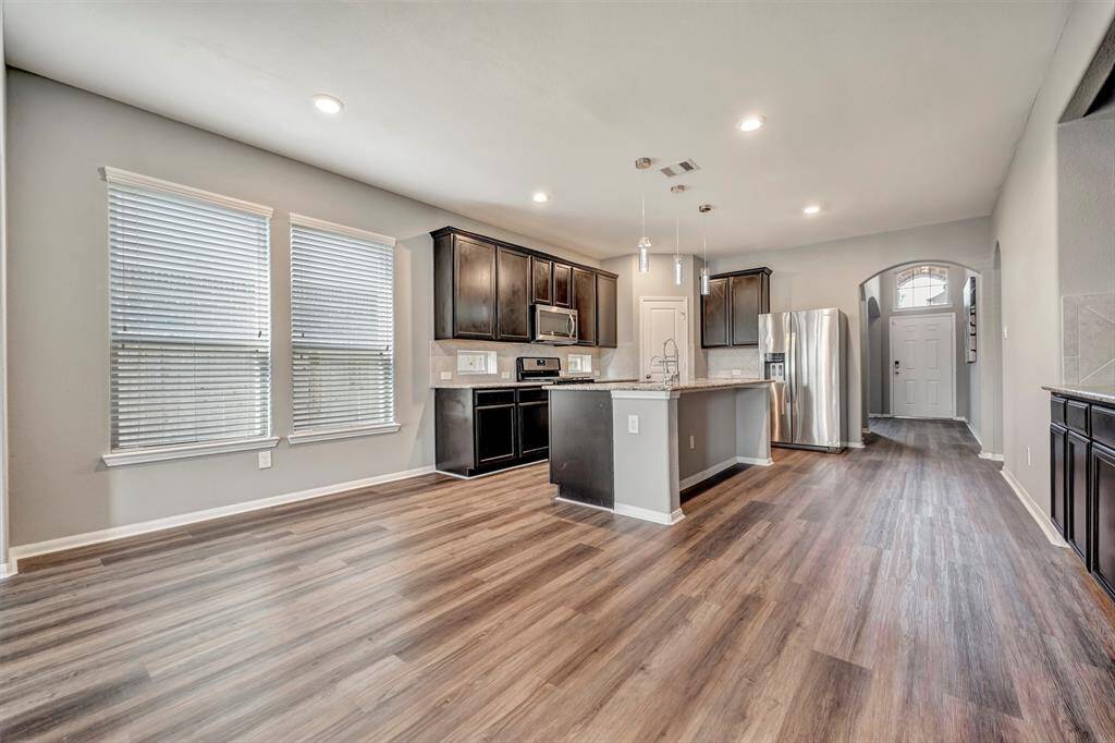 Spacious kitchen with modern appliances and dark wood cabinets, featuring a large island and ample natural light from two windows. The open layout flows into the living area with wood flooring, and an arched entryway leads to the front door.