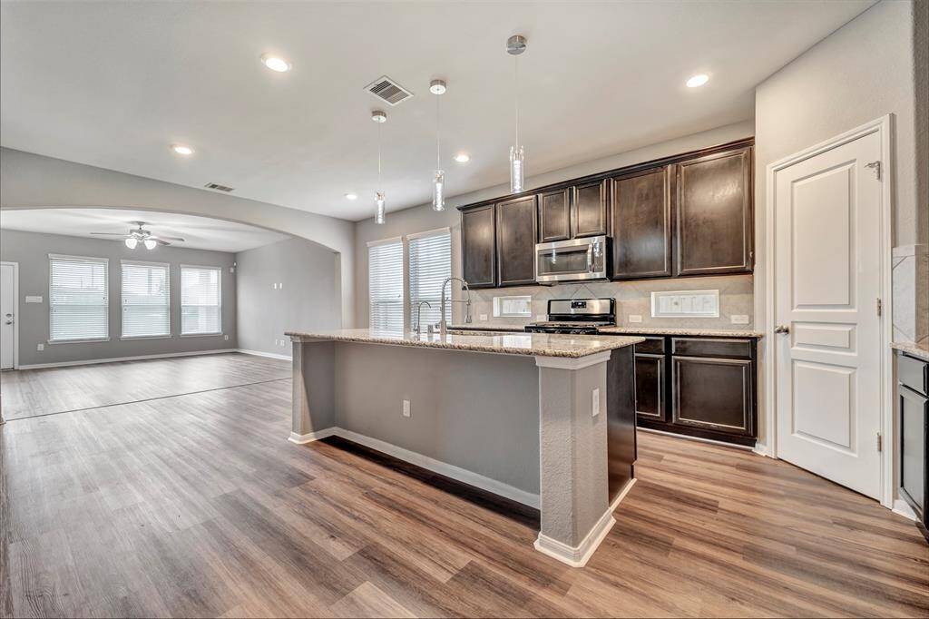 This photo showcases a modern open-concept kitchen and living area with sleek, dark cabinetry, stainless steel appliances, and an island. The space features hardwood floors, recessed lighting, and ample natural light from large windows.