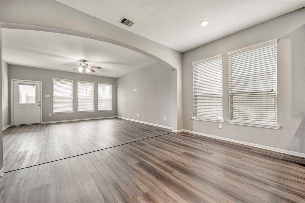 This spacious living area features sleek hardwood floors, neutral gray walls, and abundant natural light from multiple large windows. The open layout includes a ceiling fan for comfort and offers a welcoming entrance through a door on the left.