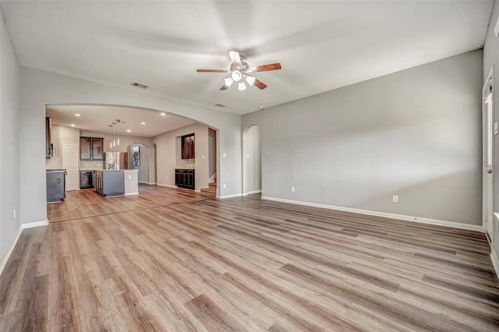 This spacious, open-concept living area features modern wood flooring, neutral walls, and a ceiling fan. It seamlessly connects to a stylish kitchen with stainless steel appliances and an island, ideal for entertaining.