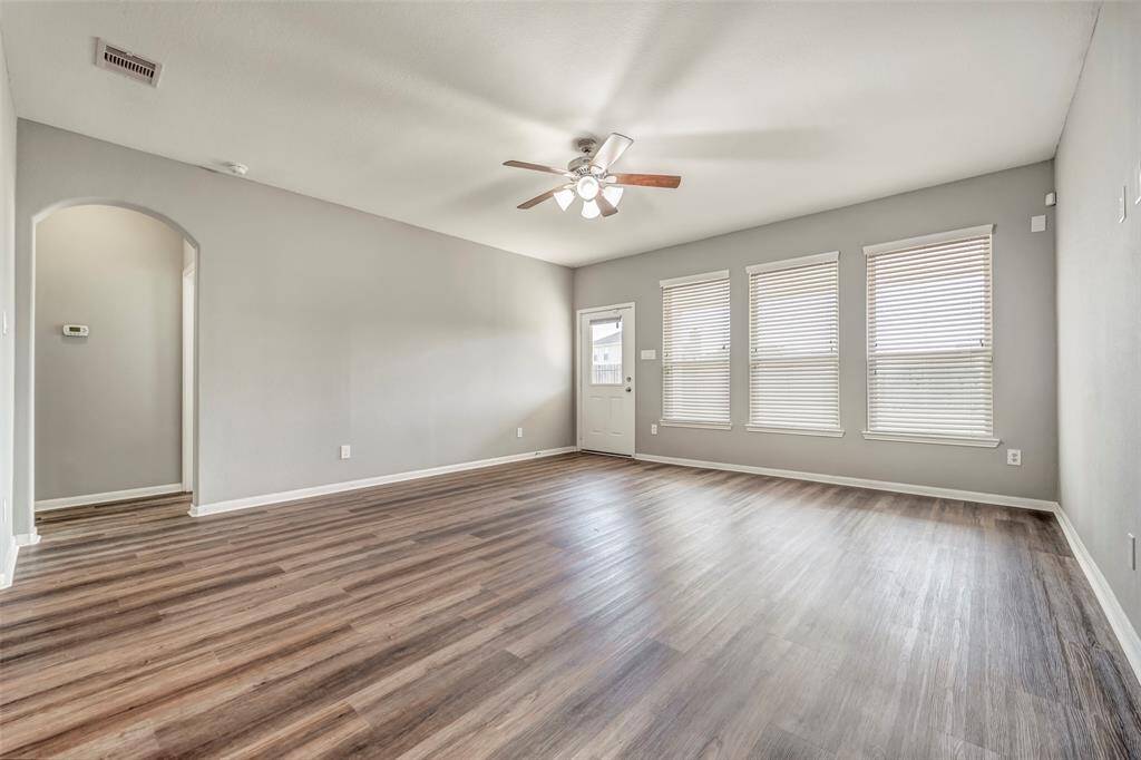 This spacious living room features a modern light gray color scheme, large windows with blinds, and stylish wood-look flooring. A ceiling fan provides comfort, and an arched doorway adds an elegant touch.