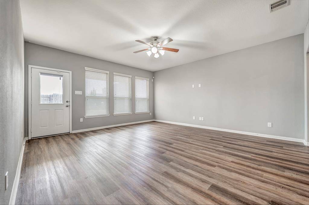 This spacious living room features modern laminate flooring, neutral gray walls, and ample natural light from multiple windows. A ceiling fan and a door leading outside complete the space.