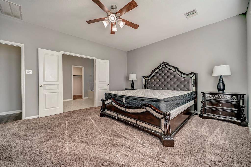 Spacious bedroom with plush carpeting, featuring a stylish dark wood bed and matching nightstands. The room is well-lit and includes a ceiling fan, with an en-suite bathroom accessible through double doors.