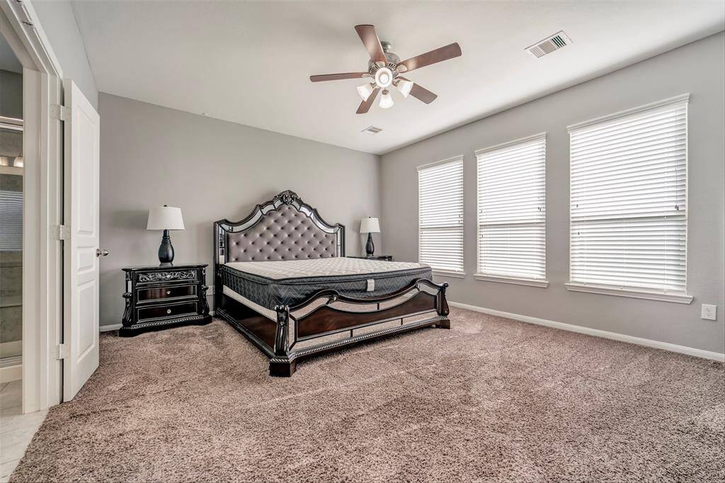 Spacious bedroom with plush carpet, neutral walls, and ample natural light from three large windows. Features a stylish ceiling fan, elegant bed, and matching nightstands, with an en-suite bathroom entrance.