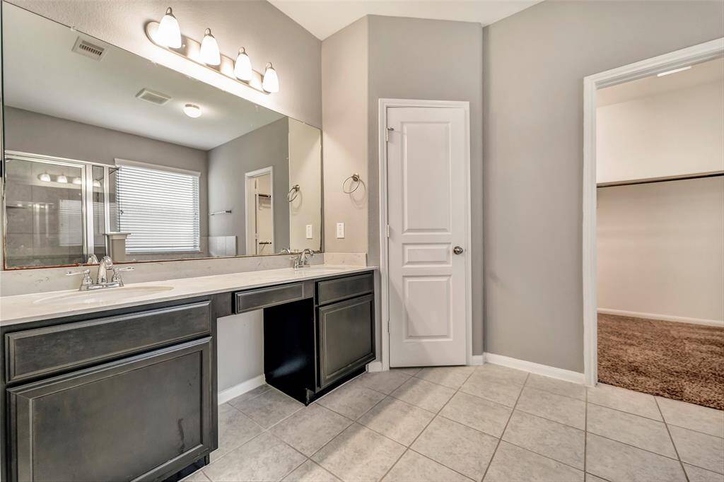 This bathroom features a spacious dual-sink vanity with dark cabinets, a large mirror, and ample lighting. It includes a walk-in closet, a tiled floor, and a shower area with natural light from a nearby window.