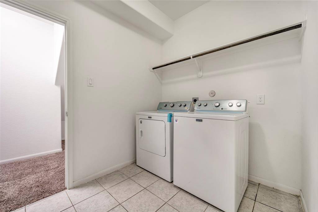This photo shows a laundry room with a washer and dryer, overhead shelving, and tiled flooring. The space is bright with a doorway leading to a carpeted area.