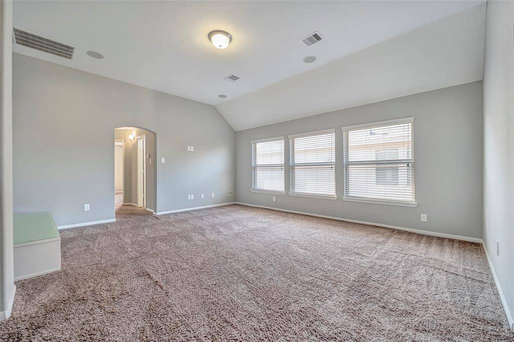 Spacious carpeted room with vaulted ceiling, large windows for natural light, and an arched doorway leading to another area. Neutral colors and ample space make it versatile for various uses.
