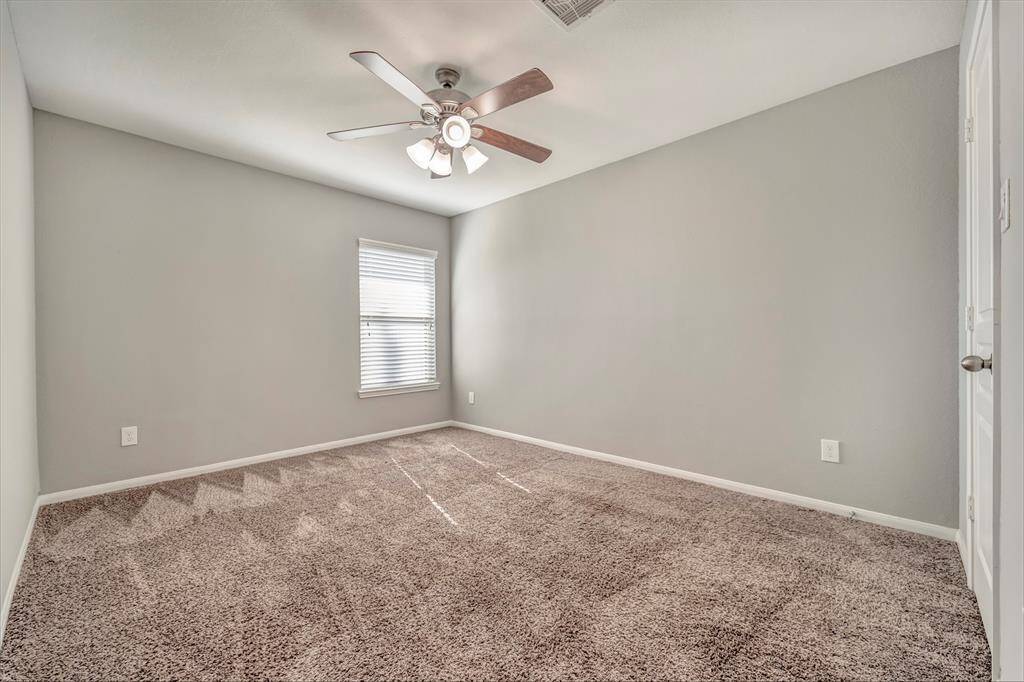 This is a bright, cozy bedroom featuring soft carpet flooring, neutral gray walls, and a ceiling fan. A window allows natural light to fill the space, creating a welcoming atmosphere.