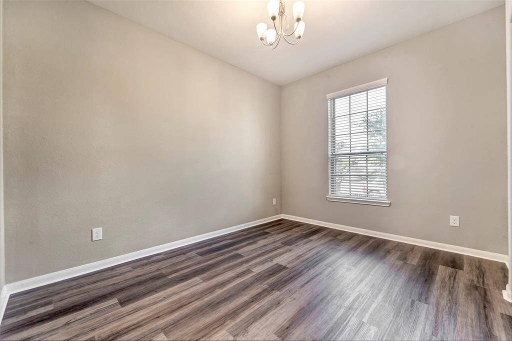 This room features a neutral color palette with freshly painted walls, wood-look flooring, and a window offering natural light. A modern chandelier adds a touch of elegance, making it a versatile space for various uses.