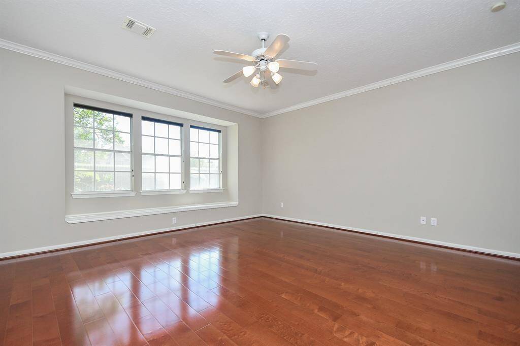 This spacious primary bedroom features gleaming hardwood floors, a large window for natural light, neutral walls, and a ceiling fan for comfort.