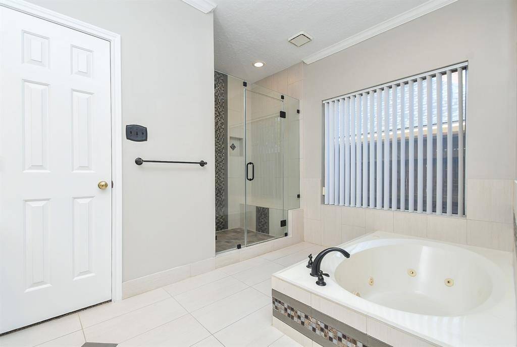 This bathroom features a spacious glass-enclosed shower and a large jetted tub. It has modern fixtures, neutral tiles, and a window with vertical blinds, providing a bright and relaxing atmosphere.