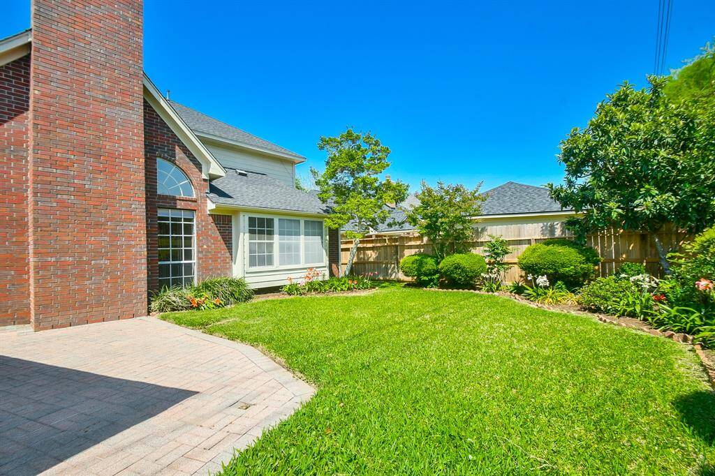 This photo showcases a charming backyard with a well-maintained lawn, surrounded by lush greenery and a variety of plants. The brick exterior of the house features large windows, allowing for plenty of natural light. A wooden fence provides privacy, creating a serene outdoor space.