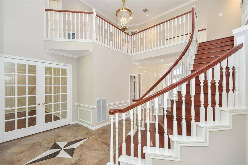 This elegant foyer features a grand curved staircase with rich wooden steps and white balusters, leading to an upper landing. The space is accented with a stylish chandelier, French doors, and a decorative tiled floor, creating an inviting entrance.