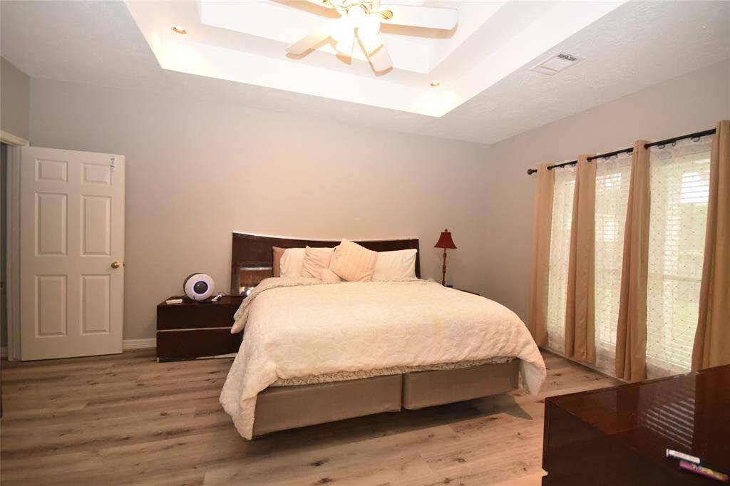 The spacious master bedroom features wood flooring and large windows that frame the lush green backyard. Recessed lighting casts a warm glow, and the stylish tray ceiling enhances the sense of openness, creating a serene and inviting retreat.