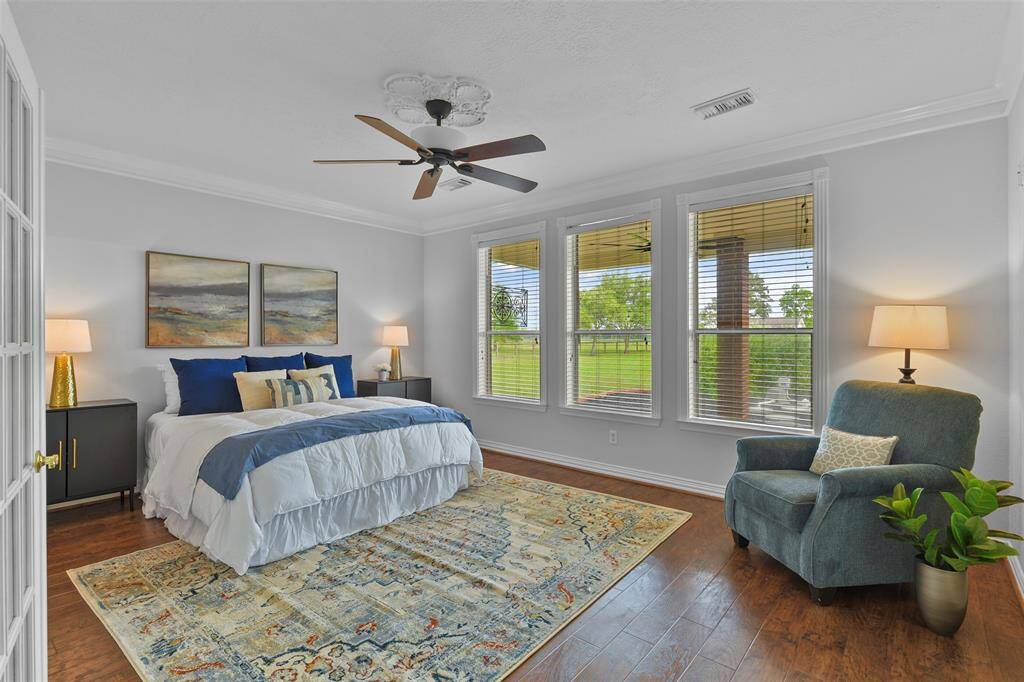 Escape to the first-floor master suite and find French doors, fresh neutral paint trimmed with crisp white crown molding, a ceiling fan with ceiling medallion, luxury vinyl plank floors, and a trio of rear-facing windows shaded by 2-inch blinds.