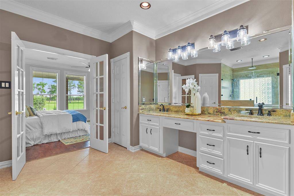 French doors open from the master suite into this en suite master bathroom with a lengthy granite countertop with dual rectangular basin sinks, oil-rubbed bronze hardware, freshly painted white cabinetry, a beveled frame mirror, and stylishly updated lighting.