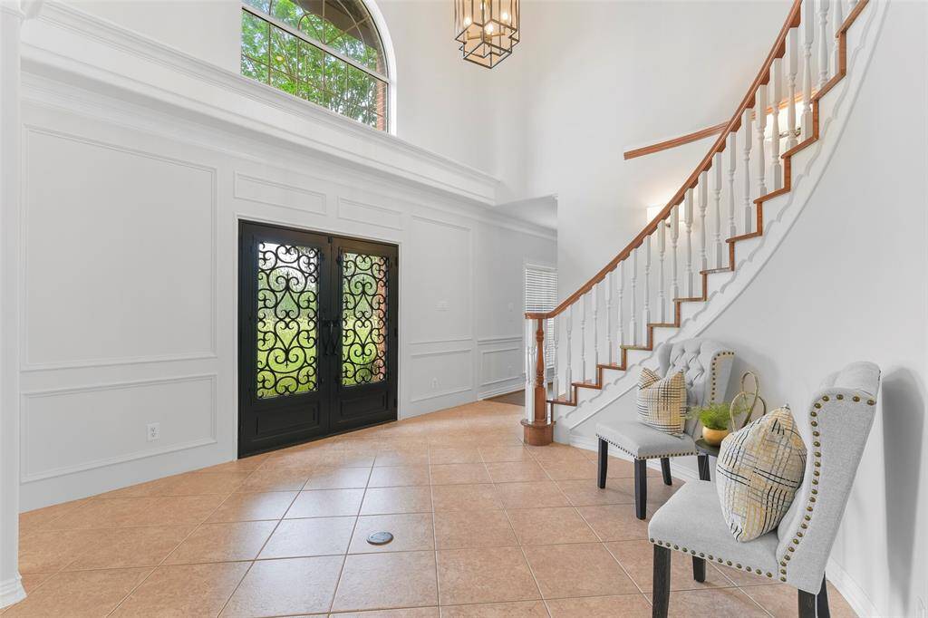 A pair of scrolled iron double front doors with rainfall privacy glass open into a grand foyer with fresh neutral paint, ornate moldings, tile floors, soaring ceilings, stylishly updated lighting, and a sweeping staircase with wood treads and handrails.
