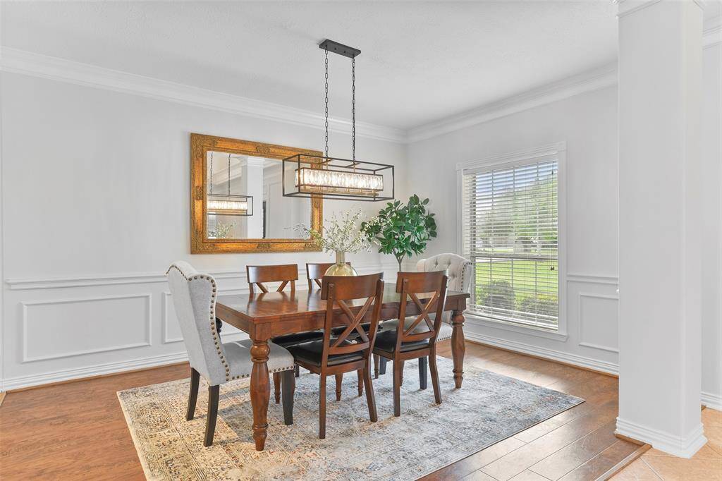 The impressive foyer makes way to an elegant formal dining room complete with wood-look luxury vinyl plank flooring, fresh neutral paint, ornate moldings, a stylish black, gold, & crystal caged chandelier, and a front-facing window shaded by 2-inch blinds.